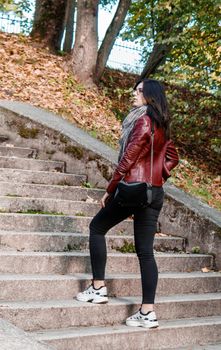 young girl in brown jacket and black jeans standing on the stairs in city park on sunny autumn day