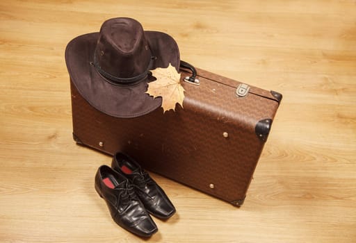 set of men's felt hat with a yellow maple leaf, black shoes and a suitcase on a wooden floor