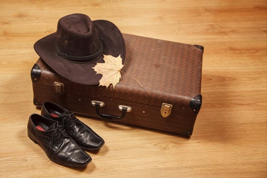 set of men's felt hat with a yellow maple leaf, black shoes and a suitcase on a wooden floor