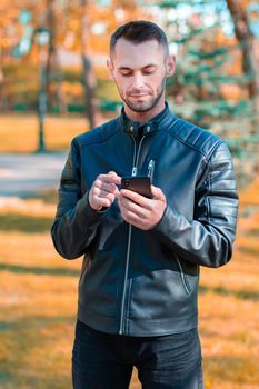 Youthful Guy Using Black Smartphone at the Beautiful Autumn Park. Handsome Young Man with Mobile Phone at Sunny Day - Medium Long Shot