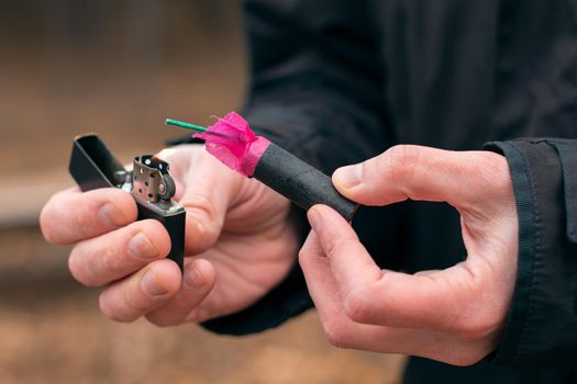 Setting Fire to the Firecracker. Man in Black Clothes Lighting Up the Petard. Firing Up the Pyrotechnics with a Black Gasoline Lighter Outdoors