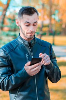 Youthful Guy Was Surprised Using Black Smartphone at the Beautiful Autumn Park. Handsome Young Man with Mobile Phone at Sunny Day - Medium Shot Portrait