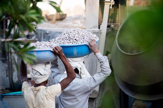 Jaipur, Rajasthan, India - circa 2020 : zoomed in shot of indian underprivleged construction workers carrying sand, cement, stones and water on their head to load into a mixer to make concrete for the making of infrastructure real estate, homes and offices in India. Shows the manual labor and hard work employed by the real estate and construction industry with lack of safety in the country of India