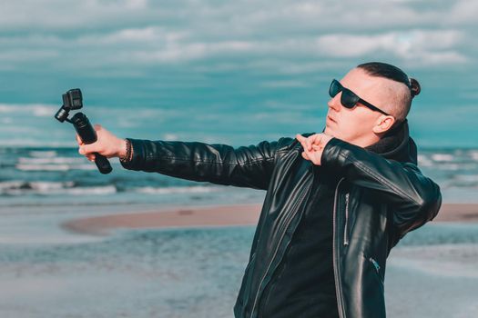 Young Blogger in Sunglasses Making Selfie or Streaming Video at the Beach Using Action Camera with Gimbal Camera Stabilizer. Hipster in Black Clothes Making Photo Against the Sea