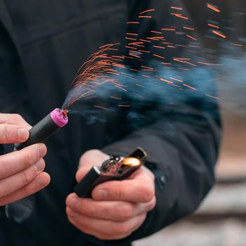 The Firecracker in a Hand. Man Holding a Burning Petard in His Hand. A Human with a Pyrotechnics that Burns with Sparks and Smoke Outdoors