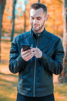 Youthful Satisfied Guy Using Black Smartphone at the Beautiful Autumn Park. Handsome Smiling Young Man with Mobile Phone at Sunny Day - Medium Shot Portrait