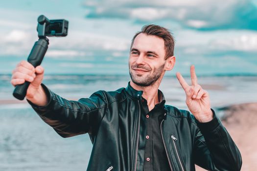 Smiling Young Blogger Making Selfie or Streaming Video at the Beach Using Action Camera with Gimbal Camera Stabilizer. Man in Black Clothes Making Photo Showing Peace