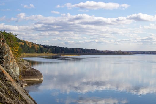 Beautiful, wide autumn river among forests and rocky shore. A calm and quiet place with autumn colors. Reflection of clouds in the water in good weather