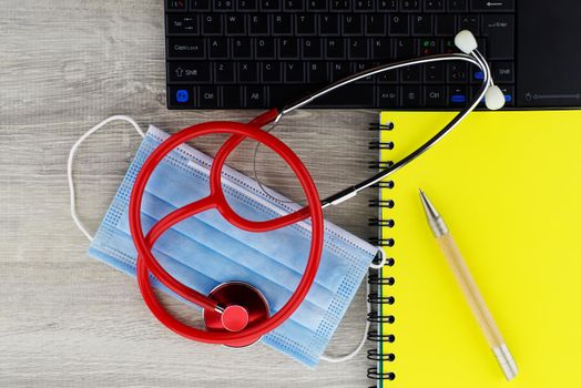 Coronavirus or Covid-19 medical concept with a colorful red stethoscope coiled over a surgical face mask alongside a notebook and pen on a wooden desk with computer keyboard viewed top down