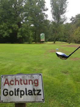 Rectangular sign with inscription in German - Caution golf course
in the background the green of a golf course with golf ball and racket. Concept Pitch and Putt