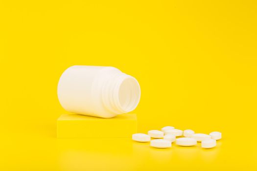 White medication bottle on podium with oval pills spilled against yellow background with copy space.Concept of wellness, healthy lifestyle, medical treatment or vitamins for kids