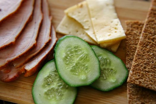 Slicing ham, cucumbers and cheese with ruddy thin breads lie on a wooden board. A healthy and healthy breakfast. A close-up of the top.