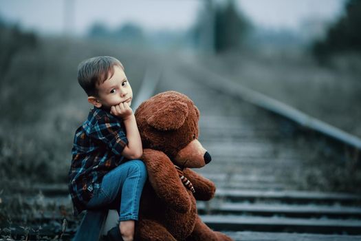 An abandoned homeless child, an orphan. A lonely boy hugs a stuffed toy and sits on the tracks, looking sadly at the camera. Street children from dysfunctional families.