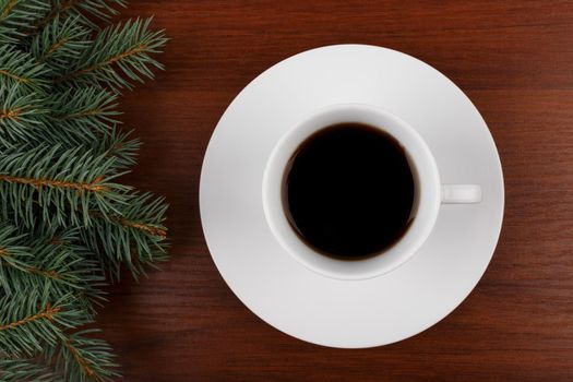 Close up of a white ceramic coffee cup with a saucer on dark shiny wooden table decorated with Christmas tree from the left side. The concept of New Year and Christmas