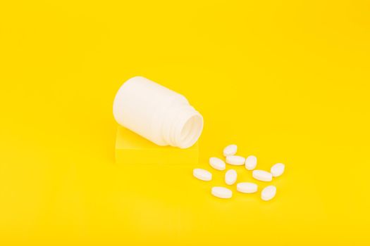 Still life with opened white medication bottle with spilled oval pills on podium against yellow background. Minimalistic concept of vitamins, minerals, supplement and healthcare