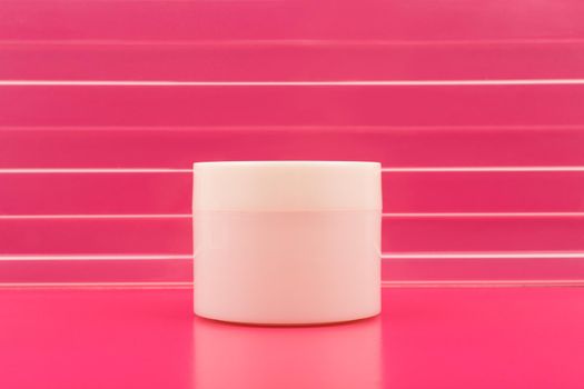 Minimalistic still life with white glossy cream jar on pink table with reflection against pink background with stripes with space for text. The concept of skin and body care and wellness
