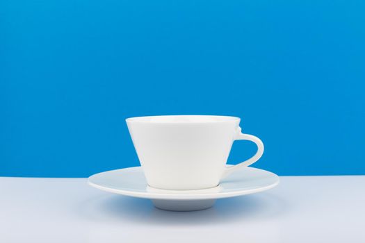 Minimalistic still life with white ceramic coffee cup with a saucer on white table against blue background with space for text