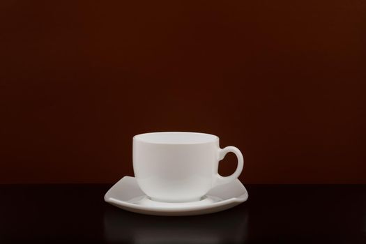 Still life with white ceramic coffee cup on plate against brown background with space for text. Concept of coffee lovers in dark tones 