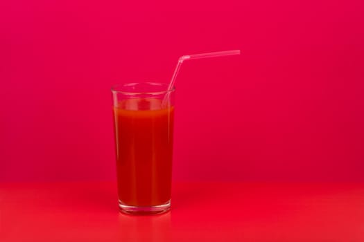 Simple still life with tomato juice in a glass with a straw against red background with a space for text. Creative concept of fresh juices and healthy lifestyle or wellbeing