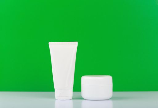 Simple still life with unbranded face and hand cream on white glossy table against green background with space for text. Concept of organic skin cosmetics or cosmetic set for hands and face 