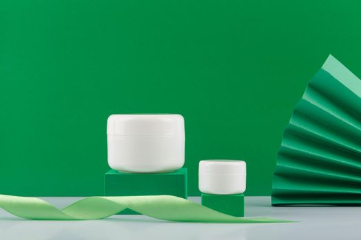 Two white glossy jars with beauty products on podiums against decorated green background. Concept of organic cosmetics with avocado, aloe vera, green tea or cucumber extract