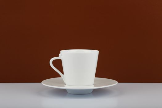 Still life with white ceramic coffee cup on plate against brown background with space for text. 