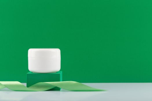 White cream, mask or balm jar on green podium with a ribbon against green background with copy space. Concept of skin cosmetics with natural ingredients like aloe vera, green tea, avocado or cucumber