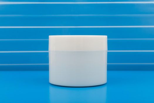 Minimalistic still life with white glossy cream jar on blue table with reflection against blue plastic background with stripes 
