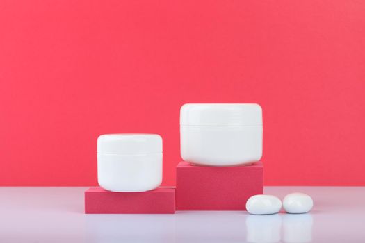 Still life with white glossy cream jars on podiums with white stones on white table against pink background with space for text. Concept of beauty. High quality photo