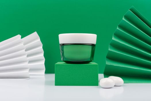 Still life with green cream jar on green podium and white glossy table against green decorated background. Luxurious organic skincare with natural ingredient for smooth, firm and young looking skin