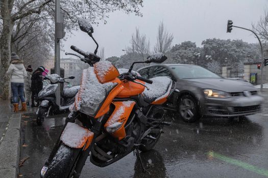 Madrid, Spain - January 07, 2021: A parked orange motorbike, covered in snow, and traffic on Avenida de Menedez Pelayo, in the middle of a wave of polar cold.