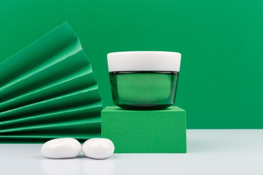Still life with green glass cream jar on green podium against green background decorared with green waver and white stones