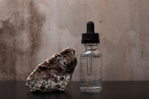 Still life with beauty serum next to a stone on black glossy table against dark marble background. Concept of luxury skincare and natural ingredients in anti aging cosmetics for firm and smooth skin