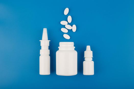 Minimalistic flat lay with white nose spray, white medication bottle with spilled pills and white eye drops on blue background. High quality photo
