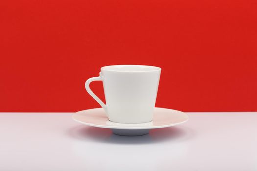 White ceramic coffee cup or tea cup on a plate on white table against red background with space for text