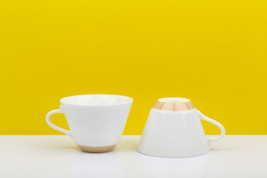 Minimalistic still life with two shiny white ceramic coffee cups on white table against bright yellow background with space for text. High quality photo