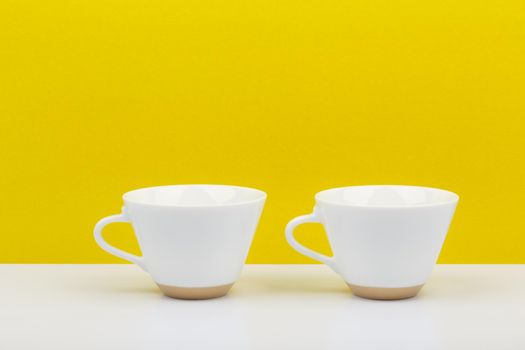 Flat lay with two white glossy ceramic cups on white table against bright yellow background with a space for text