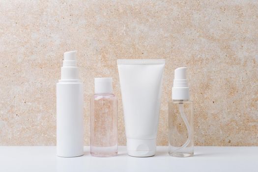 Still life with a set of cosmetic products for daily skin care on white table against beige marble background. Face cream, cleaning foam, lotion and hands cream in unbranded bottles