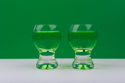 Horizontal still life with two glass shots with green sparkling drinks on white table against green background