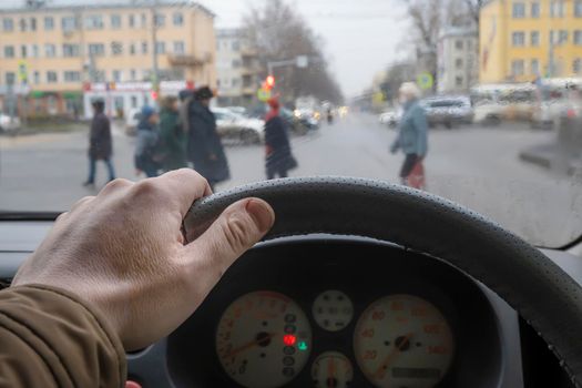 the driver's hand on the steering wheel of the car in front of the pedestrian crossing and passing pedestrians in cloudy rainy weather