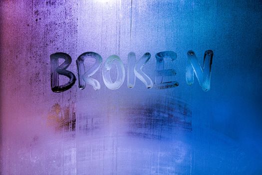 the word broken handwritten on wet window glass with selective focus and backgropund blur, neon blue color