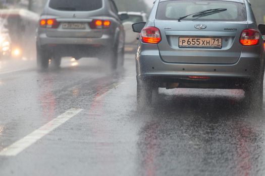 TULA, RUSSIA - JULY 14, 2020: Cars moving on asphalt road during heavy summer storm rain, back view from another car on same road. Water spraying from the wheels.