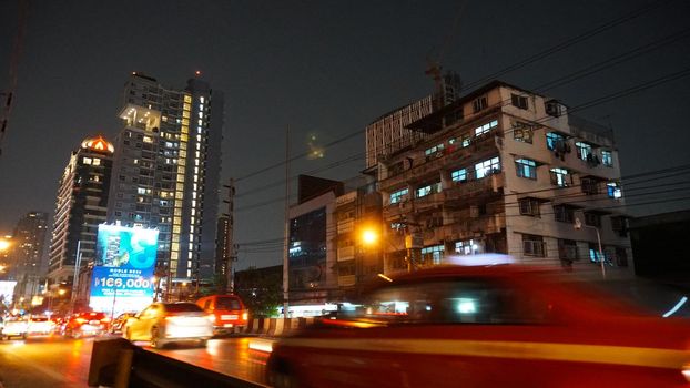 Night streets of Bangkok. Lots of cars on the road. There was a traffic jam in some places. People on motorcycles pass quickly. Everything glows. The sidewalks are almost empty. Billboards are lit.