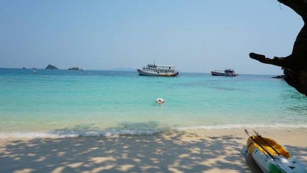 Turquoise water, clean beach and tropics. Relax. A man swims in the sea. Shadows from trees on white sand. There are ships and kayaks in the distance. Paradise lagoon. Koh Rang Island, Thailand.