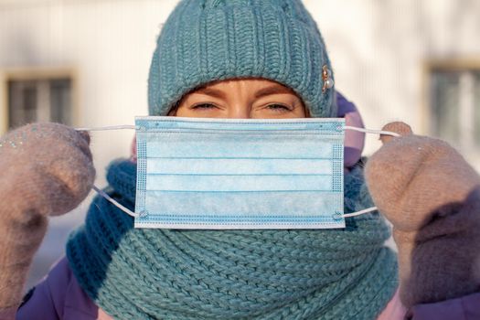 A girl in a jacket and hat in the winter on the street puts on a protective medical mask during the coronovirus pandemic. A girl holds a medical mask in her hands in the cold.