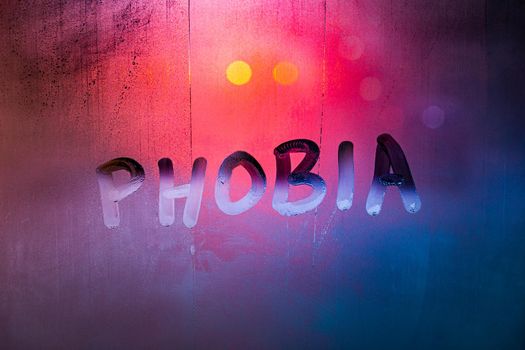 the word phobia handwritten on wet foggy window glass surface with red blue background lights.