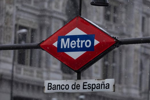 Metro Madrid sign, frozen, covered with ice and snow, in the middle of a freezing blizzard, due to the Filomena polar cold front, in the central area of Madrid, Gran Vía, Bank of Spain.