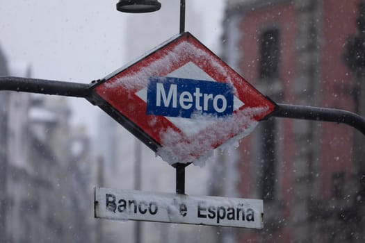 Metro Madrid sign, frozen, covered with ice and snow, in the middle of a freezing blizzard, due to the Filomena polar cold front, in the central area of Madrid, Gran Vía, Bank of Spain.