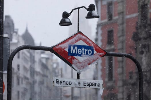 Metro Madrid sign, frozen, covered with ice and snow, in the middle of a freezing blizzard, due to the Filomena polar cold front, in the central area of Madrid, Gran Vía, Bank of Spain.