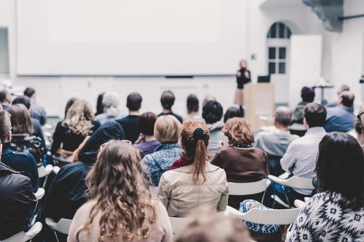 Business and entrepreneurship symposium. Female speaker giving a talk at business meeting. Audience in conference hall. Rear view of unrecognized participant in audience. Copy space on whitescreen.